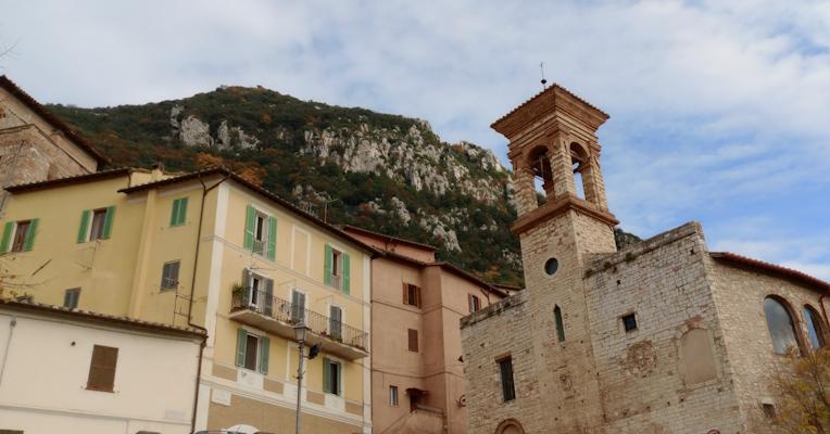 Vista di un campanile in pietra e case colorate con una montagna e cielo azzurro sullo sfondo.