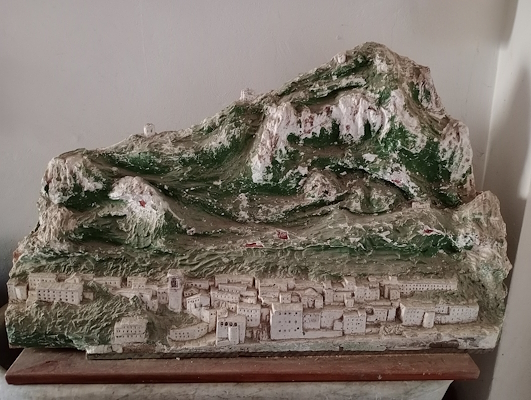 Three-dimensional model of a village at the foot of a mountain, made of painted plaster with white buildings and green reliefs.