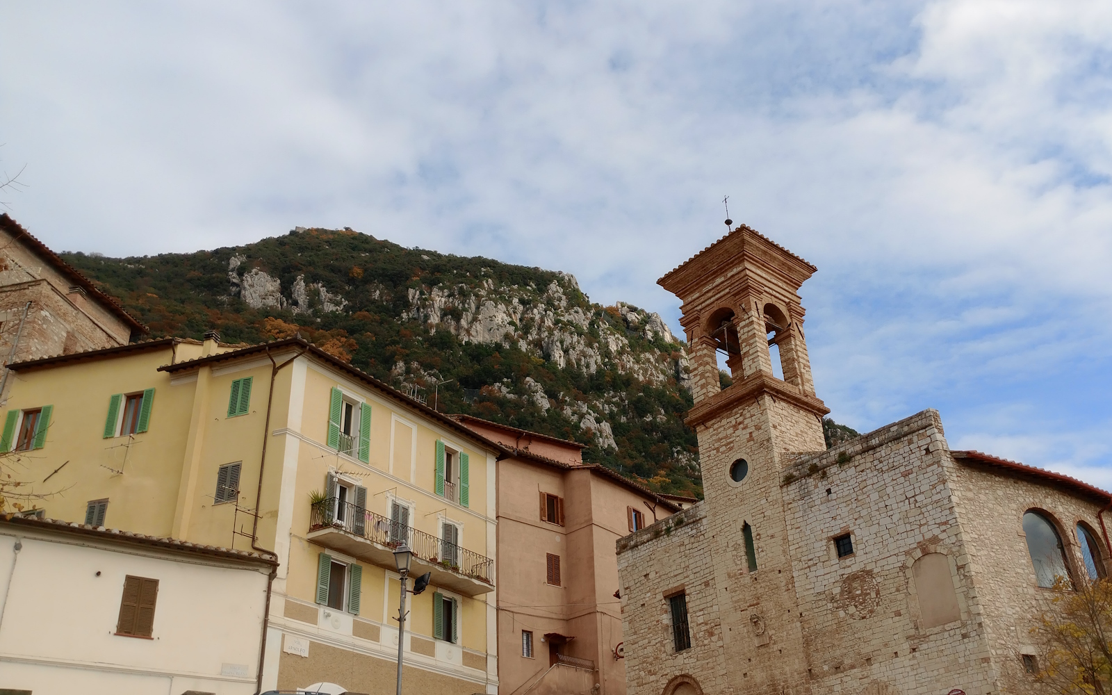 View of a stone bell tower and colorful houses with a mountain and a blue sky in the background.