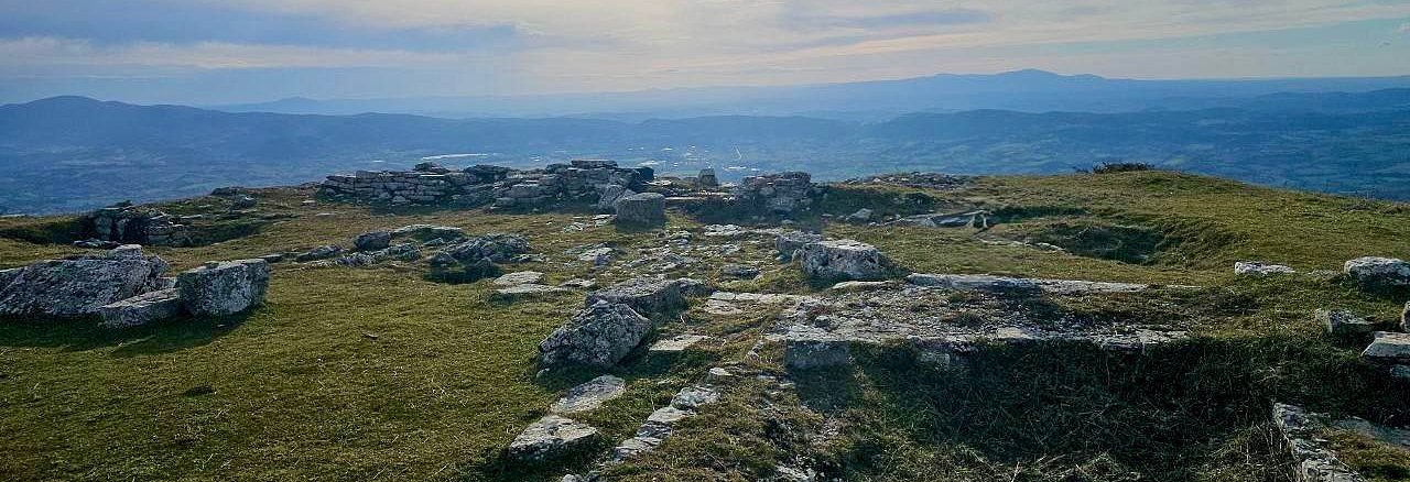 Rovine in pietra su un'altura erbosa con vista panoramica su colline e montagne all’orizzonte sotto un cielo nuvoloso.