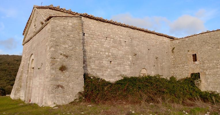 Antico edificio in pietra, circondato da vegetazione e colline, sotto un cielo azzurro con nuvole sparse.