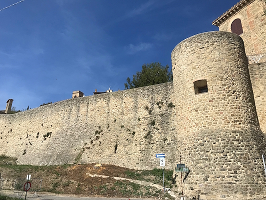 Ancient stone walls with a cylindrical tower in Gualdo Cattaneo, photographed from below under a clear sky.