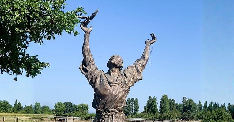 Statue of Saint Francis with arms raised and a bird on each hand, in a sunny field