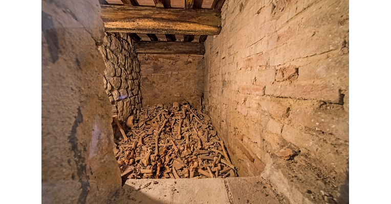 Stone-walled room with wooden ceiling containing a pile of human bones and skulls.