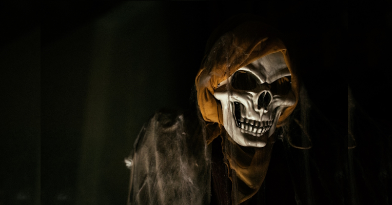  Ghostly figure with a skeleton mask and yellow hood, dimly lit in a dark setting 