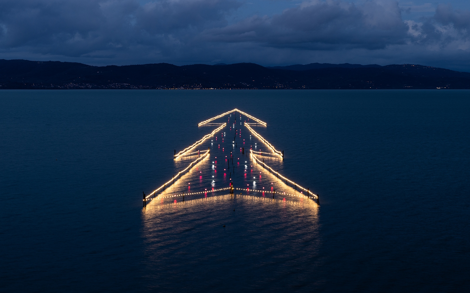 Albero di Natale illuminato galleggiante sul Lago Trasimeno al tramonto, riflesso nelle acque tranquille.