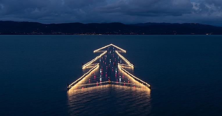 Illuminated floating Christmas Tree on Lake Trasimeno at sunset, reflected in the calm waters.