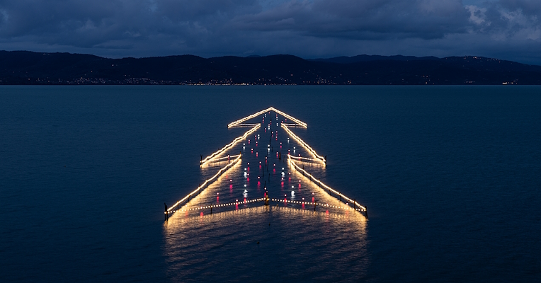 Illuminated floating Christmas Tree on Lake Trasimeno at sunset, reflected in the calm waters.