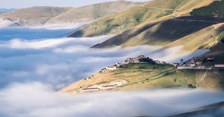 Borgo su una collina, circondato da nuvole basse tra verdi pendii e cime illuminate dalla luce del mattino.