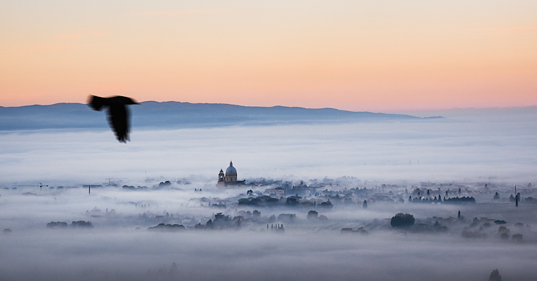 Das umbrische Tal liegt in abendlichem Nebel, im Hintergrund ragt die Kuppel der Basilika Santa Maria degli Angeli in Assisi hervor.