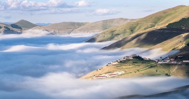 Borgo su una collina, circondato da nuvole basse tra verdi pendii e cime illuminate dalla luce del mattino.
