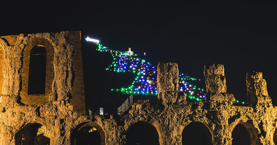  Les ruines illuminées du théâtre romain de Gubbio au premier plan, avec le sapin de Noël lumineux sur le mont Ingino, éclairé par des centaines de lumières colorées. 