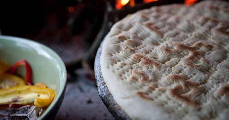 Umbrian torta al testo cooked on a testo (stone griddle) in front of a lit wood-fired oven, with grilled vegetables in a bowl.