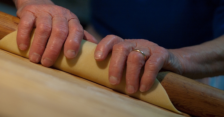 Hands rolling out a pasta sheet with a rolling pin on a floured wooden table.