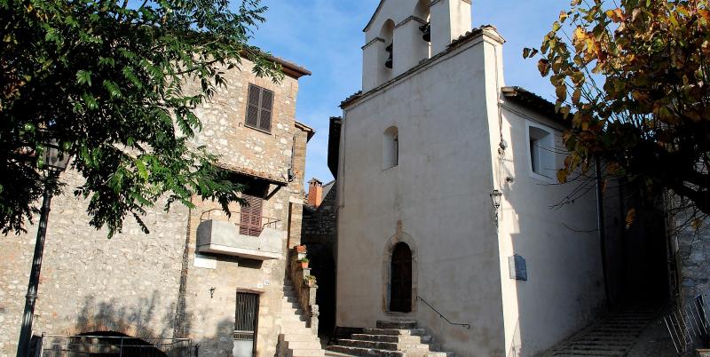 Église avec clocher-mur et escalier en pierre, jouxtée par une maison en pierre aux volets et balcon fleuri.
