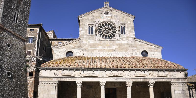 Stone façade of the Church of Santa Maria Assunta with central rose window and columned portico under blue sky.