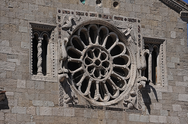 Romanesque-style rose window at the centre of the façade of the Church of Santa Maria Assunta, with sculpted decorations on the stone façade at the sides.
