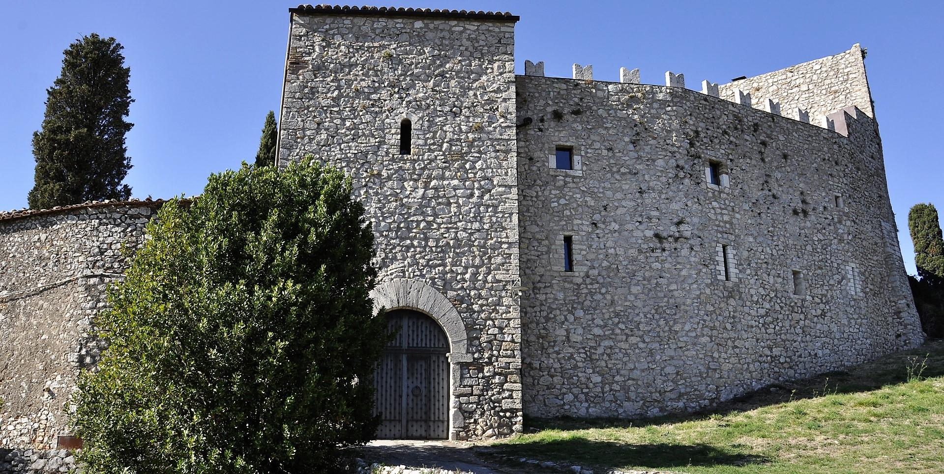 Facciata in pietra del Castello del Poggio con torre merlata, portone ad arco e mura fortificate, immerso nel verde.