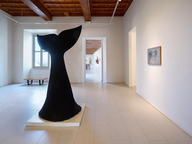 Sala espositiva con grande scultura nera al centro, opere alle pareti e soffitto ligneo a cassettoni