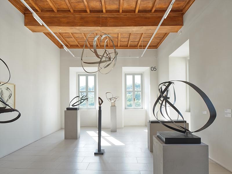 Sala espositiva con sculture astratte in metallo dalle forme curve e intrecciate, disposte su piedistalli e sospese.