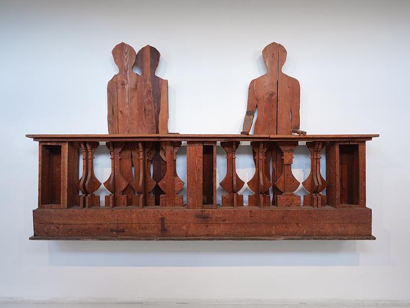 Opera in legno con balaustra e tre figure umane stilizzate, esposta su una parete bianca