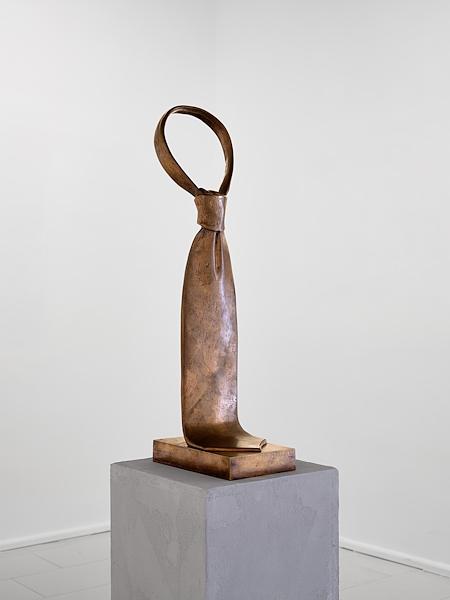 Scultura in metallo brunito raffigurante una cravatta annodata, esposta su un piedistallo.
