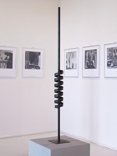 Scultura verticale in ferro nero con elemento a spirale avvolto attorno a un’asta, esposta davanti a fotografie in bianco e nero.