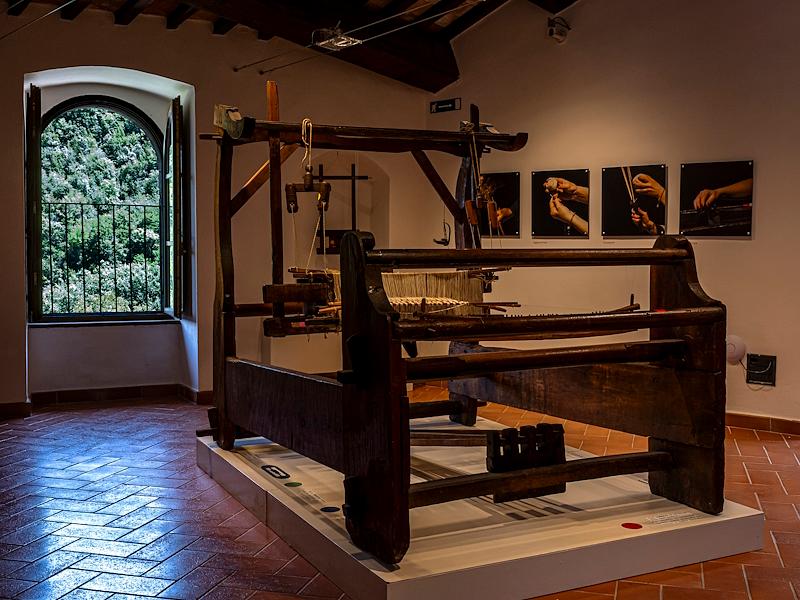 Antico telaio in legno poggiato su una pedana bianca esposto al Museo della Canapa, con fotografie alla pareti e finestra aperta