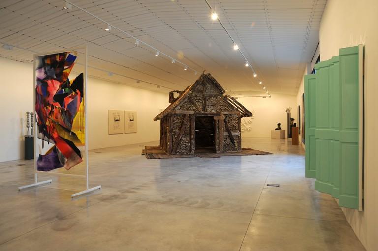 Une salle d'exposition avec une sculpture d'une cabane au centre, une œuvre abstraite à gauche et une fenêtre verte à droite.
