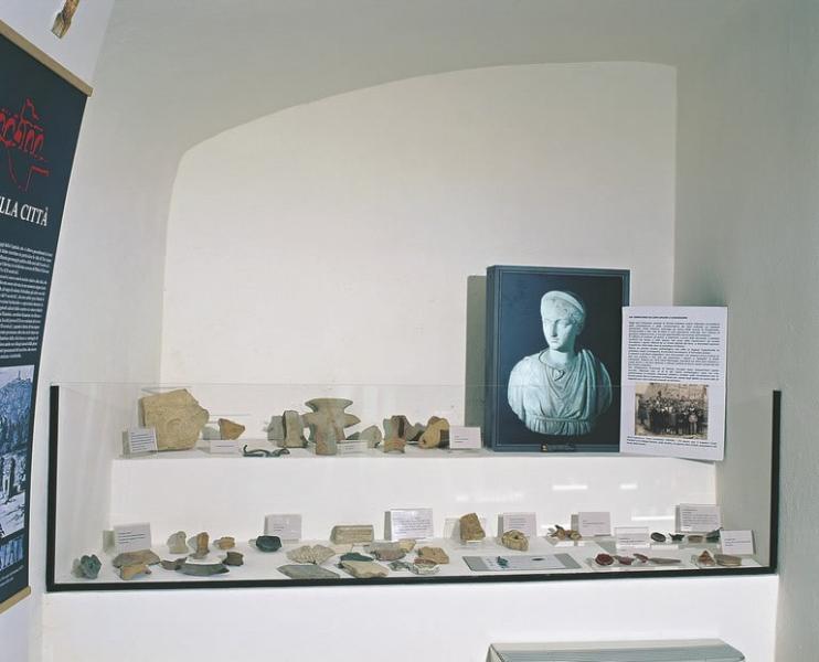 Vitrine contenant des objets en céramique, métal et pierre de l’Antiquarium d’Otricoli, à côté d’un panneau avec un buste romain.