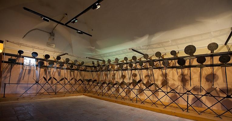 Museum room with a long row of iron wafer irons hanging on the walls, lit by soft lighting.