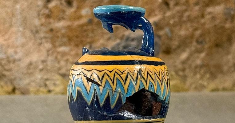 Ancien balsamaire en pâte de verre, fragmenté au niveau du col et du ventre, avec des motifs décoratifs bleus et jaunes sur fond bleu, exposé sur un support transparent