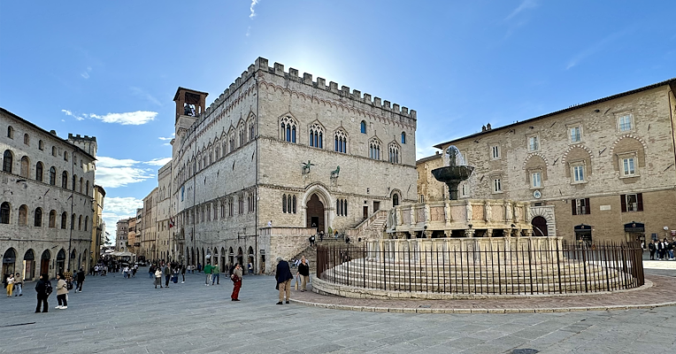 Piazza IV Novembre a Perugia con il Palazzo dei Priori e la Fontana Maggiore sotto un cielo azzurro.