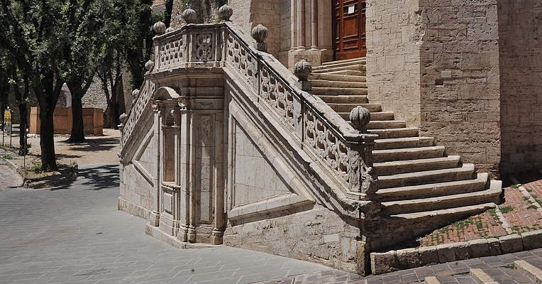 Scalinata in pietra con balaustra decorata che conduce al portale di una chiesa, tra edifici storici e un cielo azzurro.