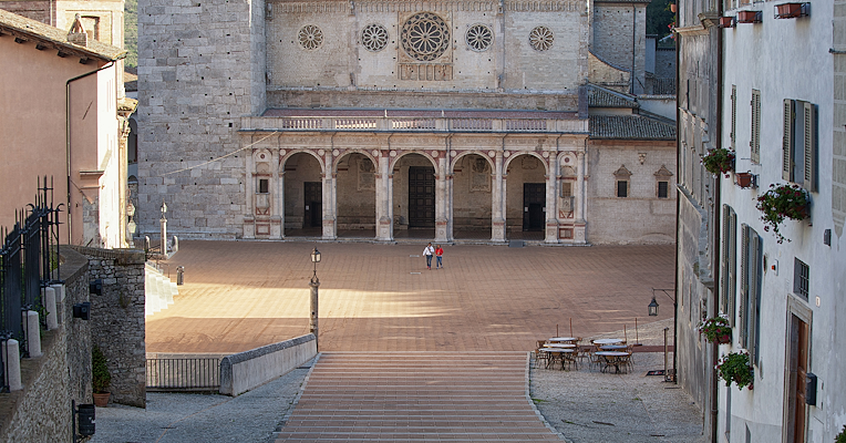 Scalinata che scende verso il Duomo di Spoleto, con facciata romanica, grande rosone e piazza ampia e luminosa.