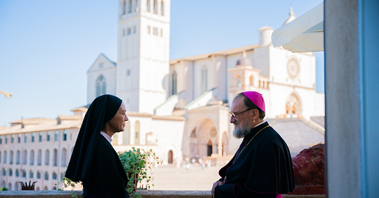 Elena Sofia Ricci, suora a sinistra, parla con un vescovo su terrazza con la Basilica di Assisi sullo sfondo.
