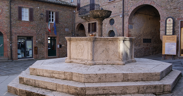 Fontana in pietra su basamento ottagonale nella piazza di Panicale, circondata da case medievali in mattoni.