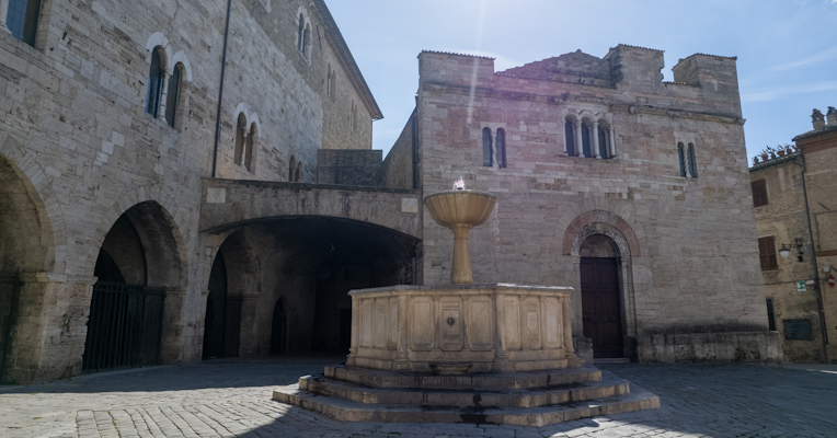 Fontana medievale in pietra nella piazza di Bevagna, circondata da palazzi storici in muratura chiara.