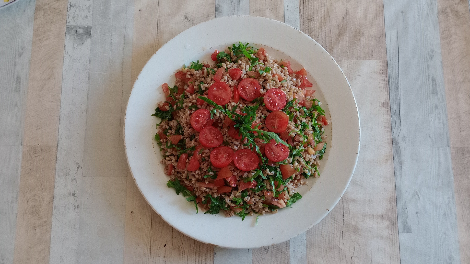 Plate of spelt salad with sliced cherry tomatoes and fresh arugula served on a light wooden table, top view