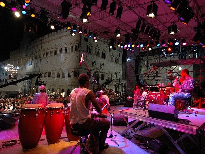 View from the stage of an Umbria Jazz concert in Perugia, with musicians playing percussion instruments in front of the audience, illuminated by colourful lights, with the Palazzo dei Priori in the background.