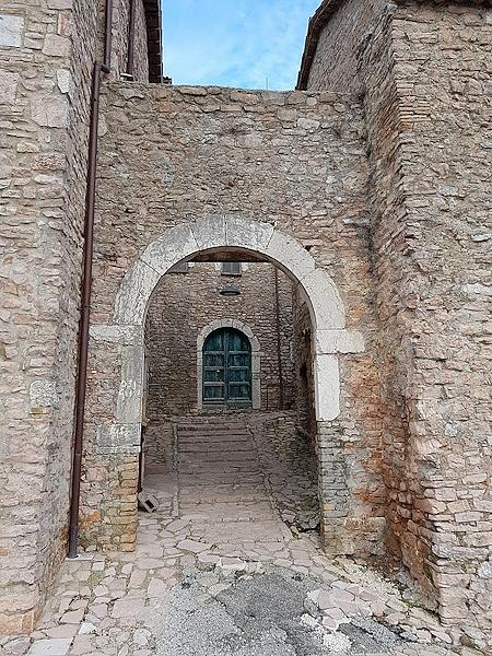 Arc en pierre à l'entrée du château de Macerino, d'où l'on aperçoit une ruelle avec des escaliers et, au fond, une porte verte
