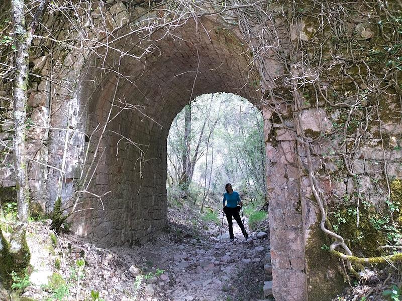 Randonneur sous un ancien pont en pierre, recouvert de mousse et de plantes grimpantes, niché dans les bois des Monts Martani, près de Macerino
