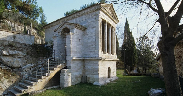Tempietto romano in pietra con colonne e scalinata, immerso nel verde e nella roccia naturale