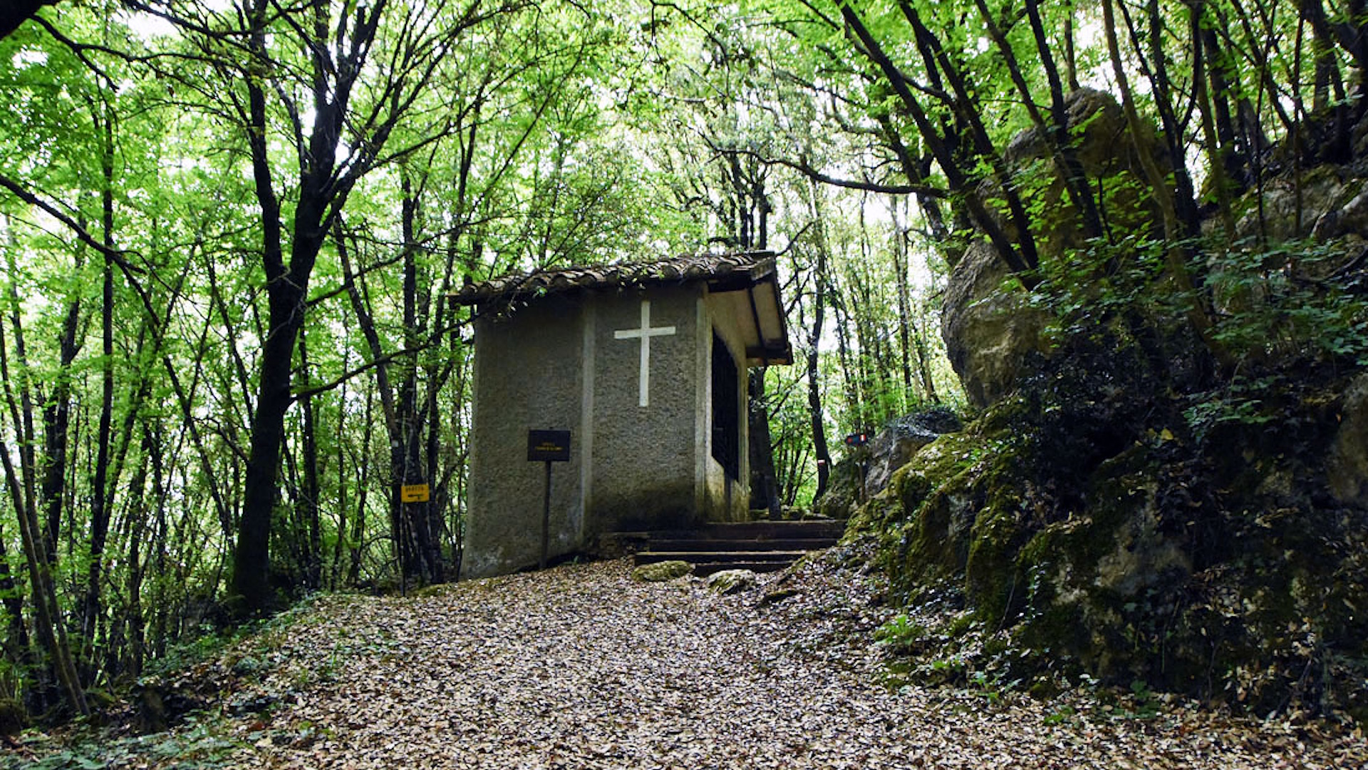 Piccola cappella dello Speco francescano immersa nel bosco, con una croce bianca sulla facciata laterale.
