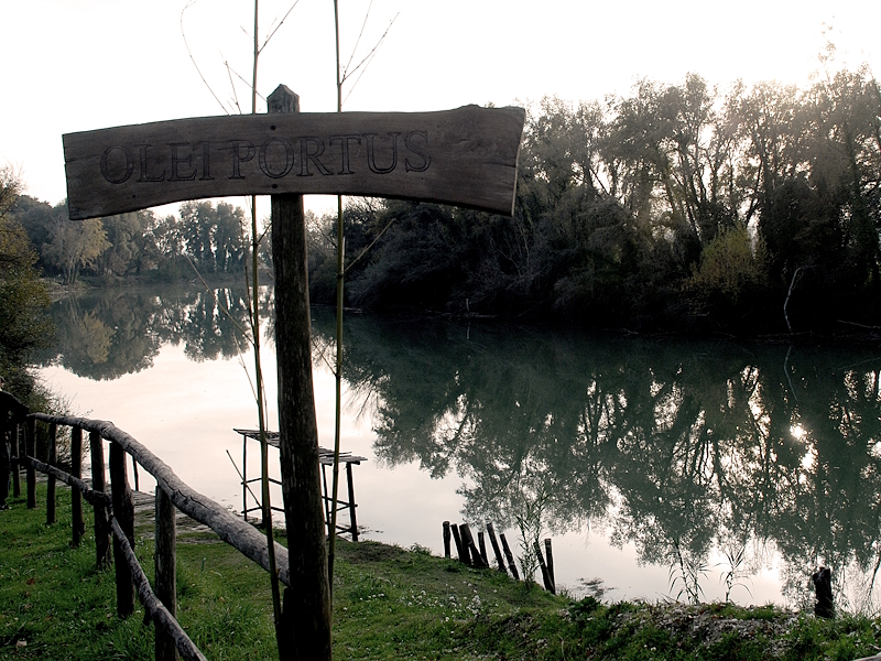 Riverbank along the Tiber at Ocriculum’s ancient river port, with the "Olei Portus" sign and riverside vegetation reflected in the calm water.