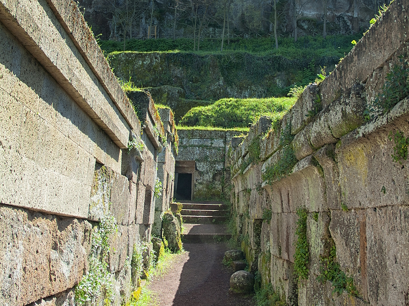 Path through the Necropolis of Crocifisso del Tufo in Orvieto, flanked by Etruscan tufa tombs, with the city’s cliff rising in the background.