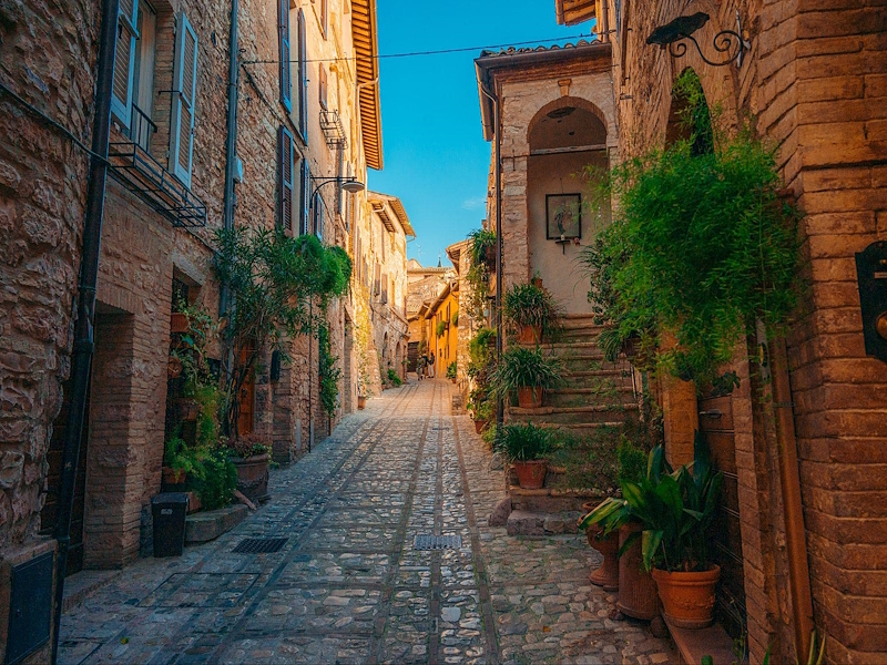 Vicolo del centro storico di Spello con case in pietra, pavimentazione in ciottoli e piante in vaso.