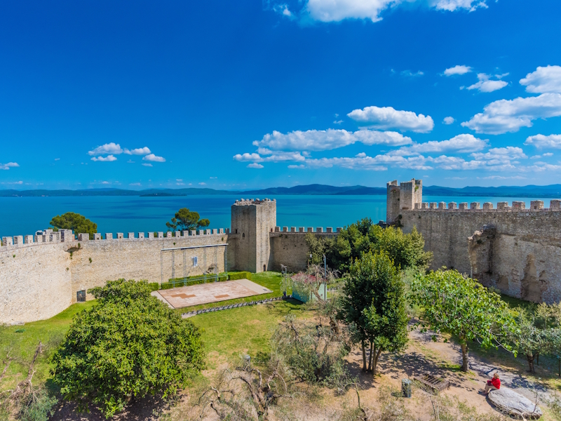 Inner courtyard of a medieval castle overlooking Lake Trasimeno, with crenellated walls and towers.