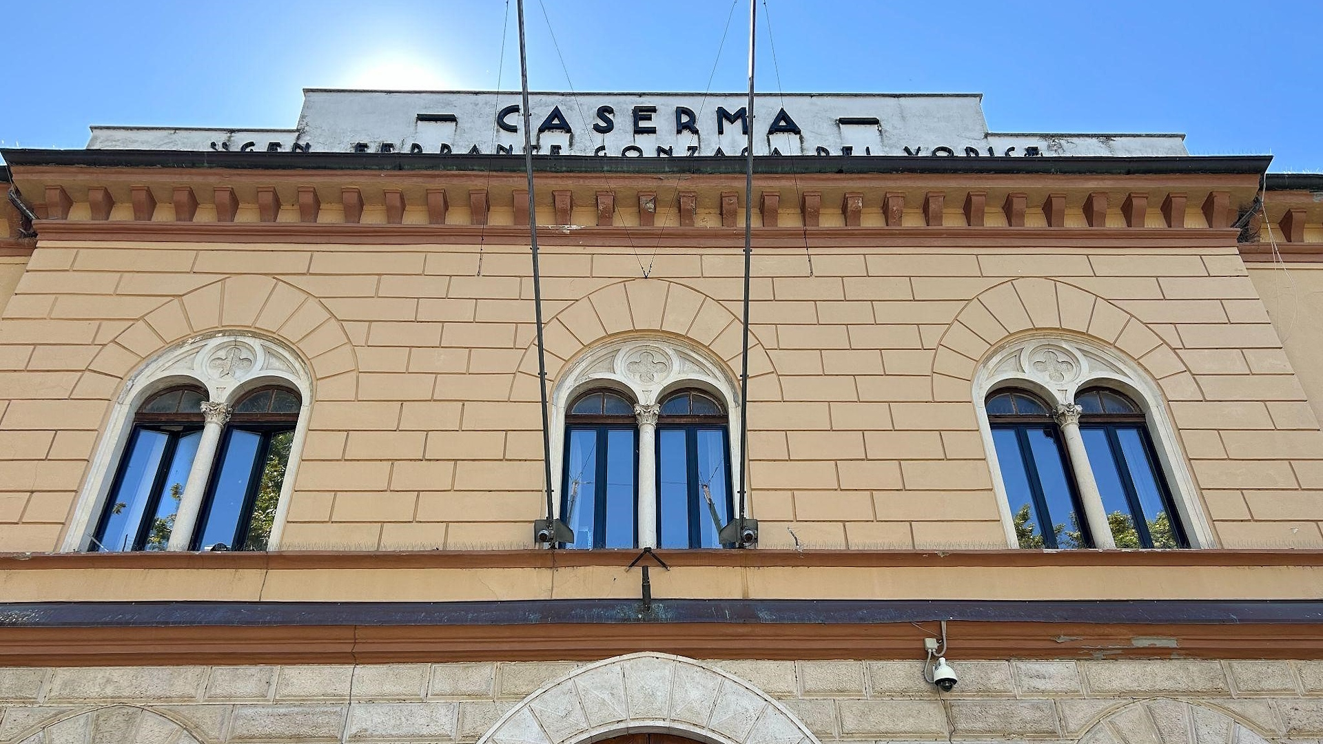 Façade of the Foligno barracks, with arched windows, a wooden entrance door and rooftop sign under a blue sky.