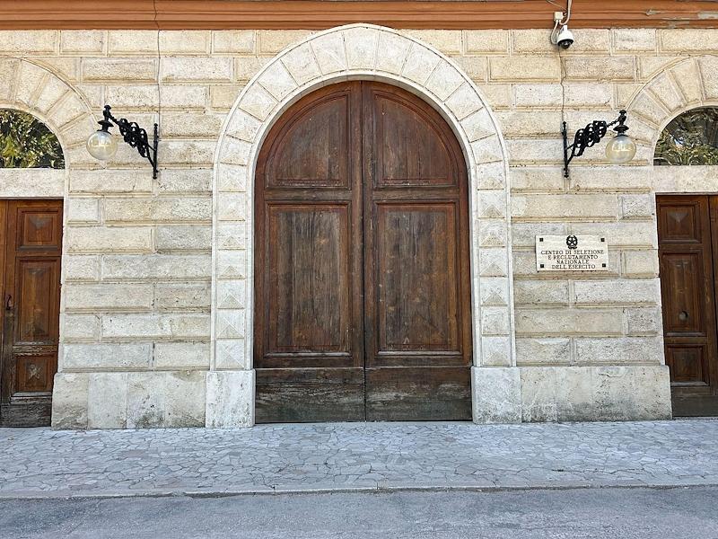 Entrance to the Foligno barracks with a large arched wooden door, stone walls and an official plaque.